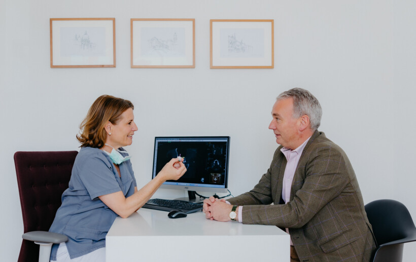 Dr. med. dent Maren Seubert erl&auml;utert einem &auml;lteren Patient seinen Eingriff im Beratungszimmer