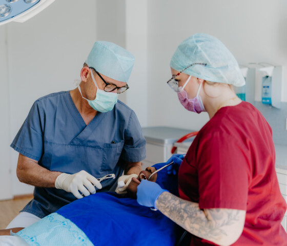 Chirurg in blauem Kasack und Assistentin in weinrotem Kasack behandeln einen Patienten unter einem blauen Tuch.
