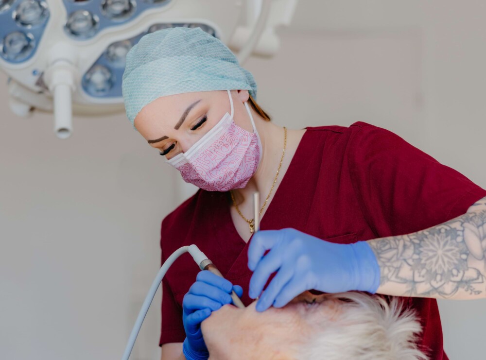 Zahnarzthelferin in weinrotem Kasack und blauen Handschuhen behandelt eine &auml;ltere Patientin.