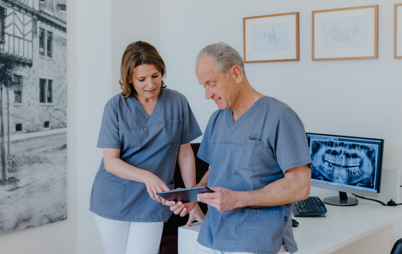 Dr. med. dent. Maren Seubert und Prof. Schierle stehen im Behandlungsraum und tauschen sich &uuml;ber eine OP-Planung aus, w&auml;hrend sie auf ein iPad blicken.