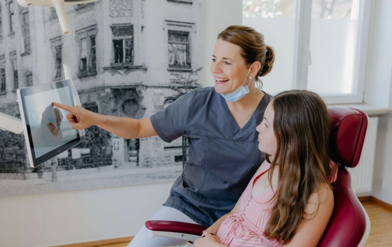 Fachzahn&auml;rztin f&uuml;r Oralchirurgie Maren Seubert erkl&auml;rt einem jungen M&auml;dchen, das auf einem Behandlungsstuhl sitzt ihre kieferorthop&auml;dische Behandlung anhand von moderner Technologie