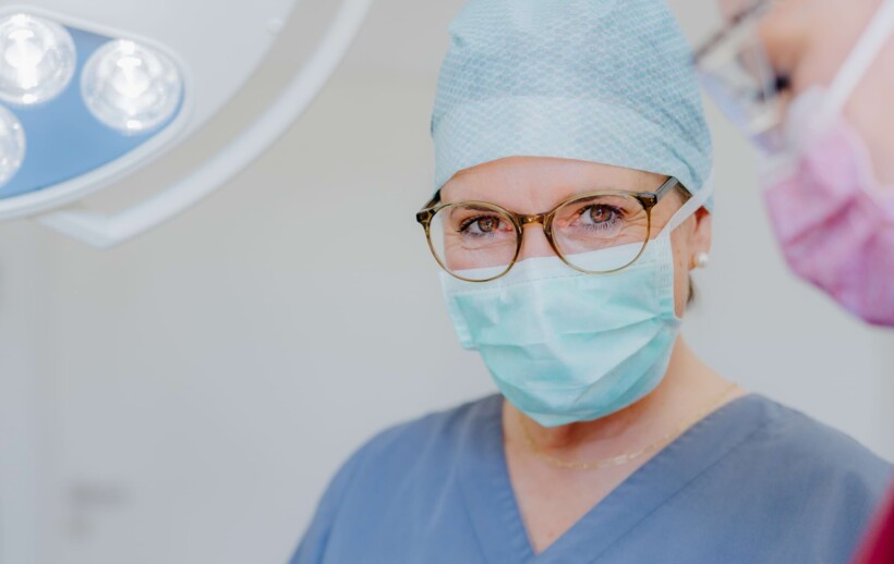 Dr. med. dent. Maren Seubert ist mir OP-Haube und Mundschutz bei einem Eingriff zu sehen und l&auml;chelt in die Kamera.