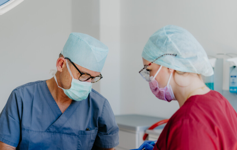 Prof. Schierle sitzt mit OP-Haube und Mundschutz einer Assistenz gegen&uuml;ber. Beide sehen konzentriert auf den Patienten zur Behandlung.