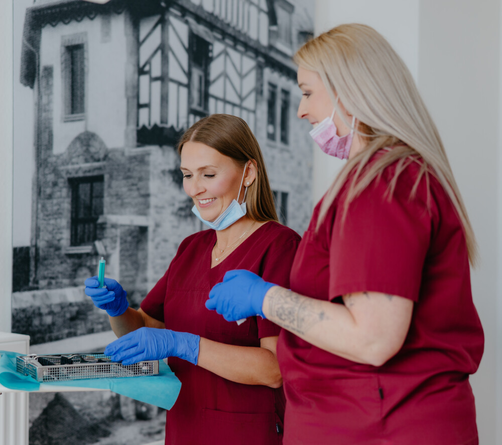 Zwei l&auml;chelnde Mitarbeiterinnen in weinroten Kasacks und blauen Handschuhen bereiten medizinische Instrumente vor.